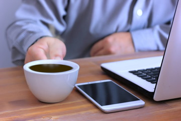 the worker holding hot coffee  with mobile phone , laptop on wood table . office man drink hot coffee before start working in the morning.