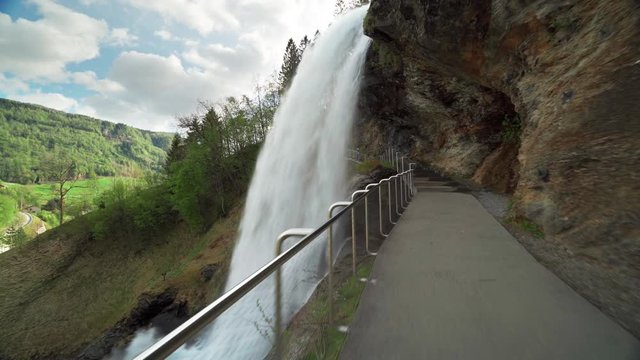 A famous Steinsdalsfossen waterfall in the village of Steine, Norway.  The wall of turbulent whitewater falling off the mountain. The pedestrian bridge is leading under the waterfall.