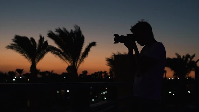 Photographer taking pictures of the Sunset Silhouette. Man photographing with a DSLR camera while the sun sets. Palmtees in the wind.