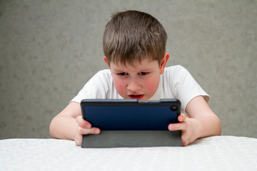 The boy emotionally plays on the digital tablet. Boy with tablet sitting on chair, on home interior background. Happy childhood. Positive emotions and energy