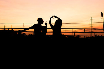Silhouette of happy couple making peace sign and heart sign over head in sunset sky evening time...
