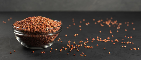 Buckwheat in a glass bowl near the copy space with spilled cereals on a black background.