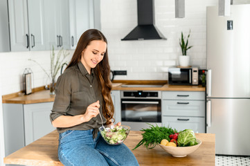 A woman in a khaki casual shirt and jeans eats a fresh wholesome salad in the kitchen from a bowl. Healthy eating concept