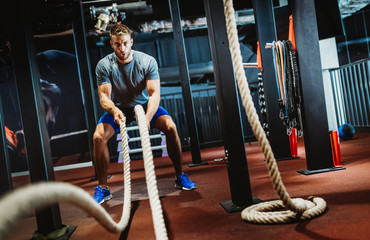Fit man working out with battle ropes at fitness gym