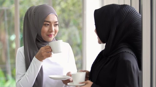 Two Asian Muslim women standing and talking in the office with a cup of coffee. Modern Muslim lifestyle concept. Footage has been recorded in 4K resolution.