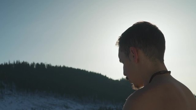 Close up view behind man breathing heavily while sitting outside in cold mountain environment with no shirt. Young guy hyperventilating to prepare to swim in icy remote wilderness lake.