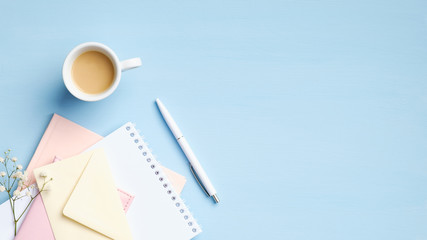 Feminine workspace with paper notebook, envelope, flowers, cup of coffee on blue background....