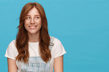 Indoor shot of young pretty redhead woman with natural makeup looking wonderingly aside with pleasant smile, dressed in casual wear while standing over blue background