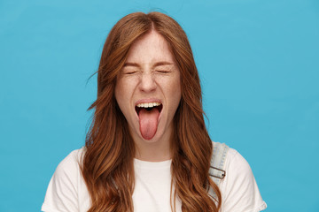 Funny shot ot young joyful redhead lady with natural makeup fooling while posing over blue background, frowning face and sticking out her tongue with closed eyes