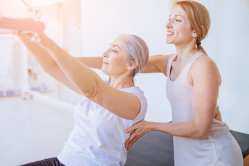 Fototapeta premium Blond female instructor helping, consulting old age grey hair beautiful woman workout pilates on reformer practice in pilates studio, working out indoor, correcting beginners, frehabilitation concept.