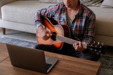 Obraz premium Music college hipster student in checkered shirt practicing acoustic guitar exercise, reading notes from laptop. Man taking an online musical courses at home during quarantine. Background, close up