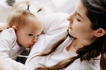 young pretty woman breastfeeding her little daughter sitting on the window sill of her bedroom