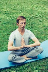 Naklejka na ściany i meble Young attractive man in sport clothes is meditating cupped in a meditation or prayer gesture with a pacified face in the park against the background of green grass, eyes closed, face with a smile.