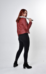 Portrait of a pretty girl with red hair wearing black jeans and boots with leather jacket.  full length standing pose holding knife a studio background.