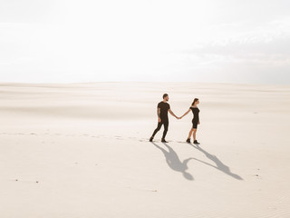 Fototapeta na wymiar Lovely attractive couple on the white sand beach or in the desert or in the sand dunes. Beach honeymoon couple holding hands walking on white sand beach 