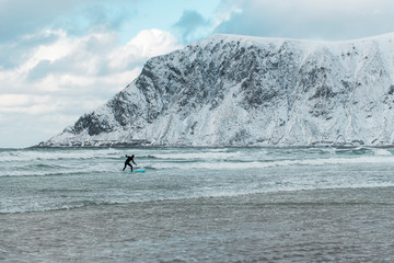 Surfing in cold ocean in Lofoten islands. Famous destination for surfers all around the world. Surfing between snowy mountains and arctic beaches. Surfers riding the waves in Norway.