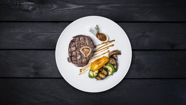 Steak osso buko with grilled vegetables rotating on a plate. On black wooden background. Top view