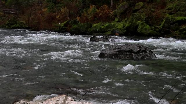 Left to right pan then panning down to rest on granite rocks framing the fast moving wild and scenic river with rich green moss background.