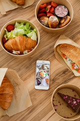 Different takeout food on wooden kitchen table. Italian panini sandwich, french croissant with salmon, pancakes, chocolate cheesecake, phone w/ food photo. Close up, top view, copy space, background.