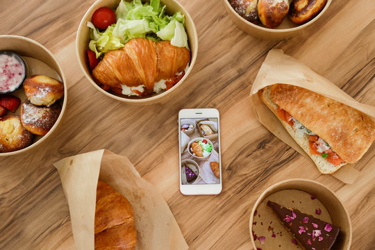 Different takeout food on wooden kitchen table. Italian panini sandwich, french croissant with salmon, pancakes, chocolate cheesecake, phone w/ food photo. Close up, top view, copy space, background.