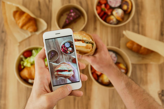Food blogger reviewing takeout food, capturing video on his phone. Italian panini sandwich, french croissant with salmon & chocolate cheesecake. Close up, top view, pov, copy space, wooden background.