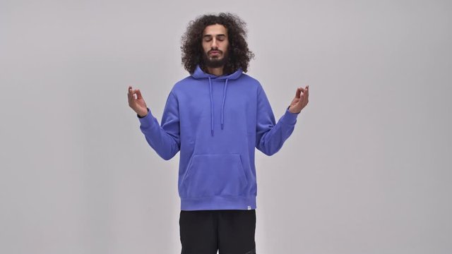 A calm young bearded brunette man with long curly hair is doing a dzen gesture with his hands over the gray wall in the studio