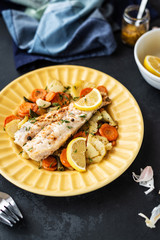 Baked hake fish in the oven with vegetables on a yellow plate on a black background. Delicious hot snacks for foodies. Selective focus