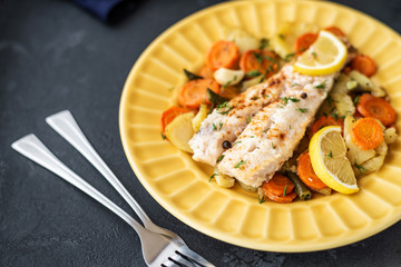 Baked hake fish in the oven with vegetables on a yellow plate on a black background. Delicious hot snacks for foodies. Selective focus