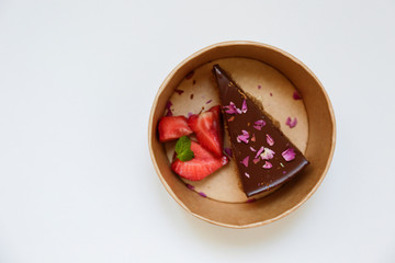 Slice of dark tripple chocolate mousse cake with rose petals decoration, strawberry and mint leaf inside of paper takeout bowl. Close up, top view, flat lay, isolated, copy space for text.