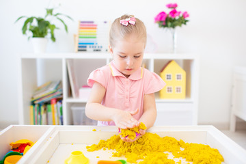Little blonde girl playing with yellow magic sand on a white table in a light room. Sensory...