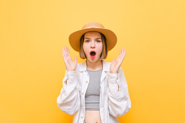 Shocked lady in hat and light clothing isolated on yellow background, looks into camera with mouth open, shouts WOW and raises palms to face. Portrait of surprised girl on orange background.