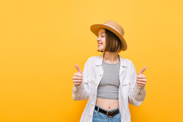 Pretty girl in stylish summer clothes and hat stands on yellow background, looks away at copy space with a smile on her face and shows thumb up gesture. Lady shows gesture like, isolated