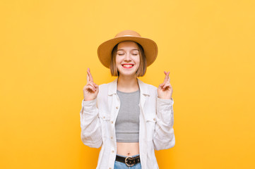 Happy girl in light clothing and hat stands on a yellow background and makes a wish with crossed...