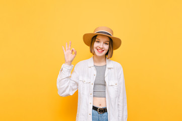Happy girl in a hat, shirt and top stands on a yellow background, shows Okay gesture and smiles, looking into the camera. Cheerful lady in summer clothes shows the OK gesture and laughs.