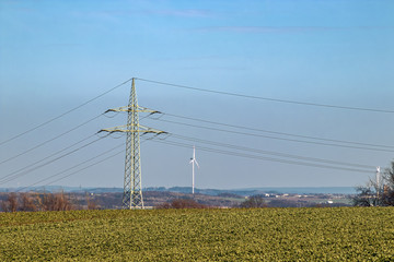 Strommast, Hohspannungsleitung, auf Feld im Frühjahr und Windkraftanlage (Windturbine) am Horizont