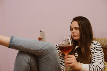 Sad young woman is using phone with glass of wine at home on sofa indoor during home isolation quarantine