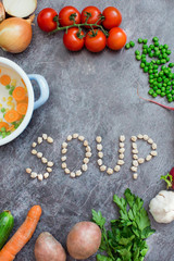 Composition with fresh vegetables on grey background, space for text. Pot with vegetables and word "soup" made of chickpea