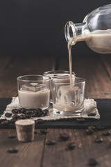 Pouring  Cream coffee liqueur in glasses with ice, surrounded by coffee beans on a dark wooden background.