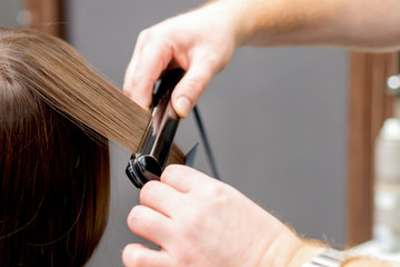 Hairdresser hands are straightening hair of woman with straightener tool in hair salon, back view.