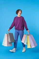 Pleasant looking young pretty dark haired curly woman walking with paper bags while shopping and smiling cheerfully, isolated over blue background