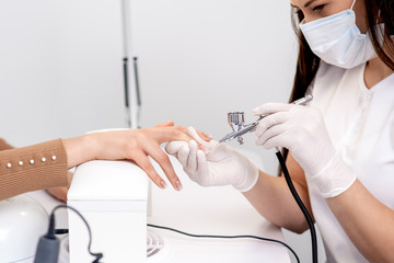 Manicure master is painting finger nails in beige color using airbrush. Woman getting fingernail...