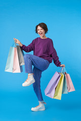 Overjoyed young lovely dark haired female with casual hairstyle being in high spirit while buying new clothes, looking excitedly at camera while posing over blue background