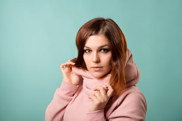 Red-haired girl with make-up and hairstyle holds hands by the pink collar of a sweatshirt against a blue background close-up.