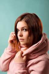 Red-haired girl with smokey eyes make-up is holding with her hands a collar of pink sweatshirt on a blue background close-up.