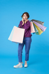 Excited young attractive short haired curly female hearing unexpected news while having phone talk, holding shopping bags while posing over blue background