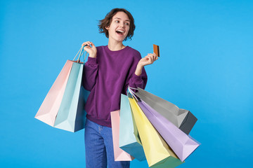 Fototapeta na wymiar Good looking young happy short haired brunette female with casual hairstyle rejoicing while making shopping with her credit card, isolated over blue background