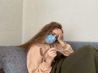 girl in a disposable medical mask is sitting at home with a headache. The girl has a high...