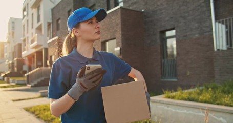 Caucasian young pretty woman, delivery worker in blue cap walking the street and carrying carton box while using smartphone, looking for route. Female courier with parcel tapping and texting on phone. - Powered by Adobe