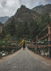 A girl in a yellow raincoat and with a backpack stands on a bridge over a mountain river and looks at the mountains