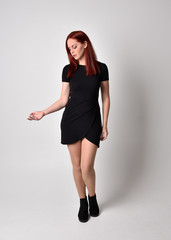 Portrait of a pretty girl with red hair wearing short black dress and boots, full length standing...